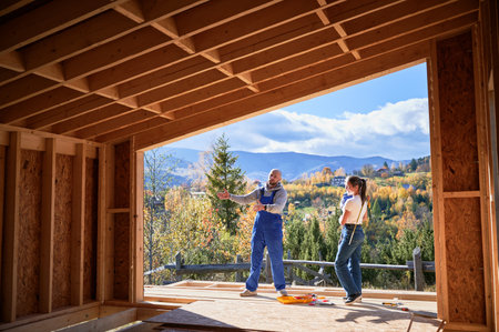 Father, mother and son building wooden frame house. Male designer in blue overalls showing future home, standing on terrace on construction site. Carpentry, family concept.の写真素材