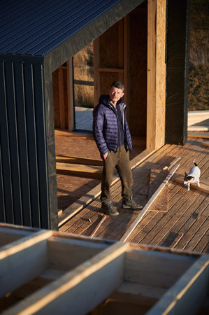 Male designer building wooden frame house. Man standing on terrace at construction site, inspecting quality of work in the evening.の写真素材