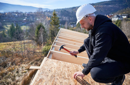 Carpenter hammering nail into OSB panel on the roof top of future cottage. Man worker building wooden frame house. Carpentry and construction concept.の写真素材