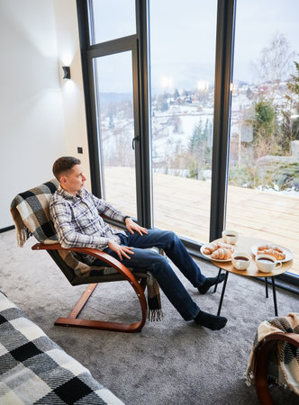 Young man enjoying weekends inside contemporary barn house. Happy male tourist sitting on chair, enjoying view of mountain landscape through panoramic windows in new cottage.の写真素材