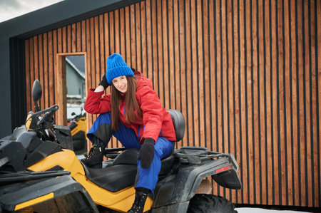 Portrait of beautiful woman posing on offroad four-wheeler ATV with wooden house on background. Concept of active leisure and winter activities.の写真素材