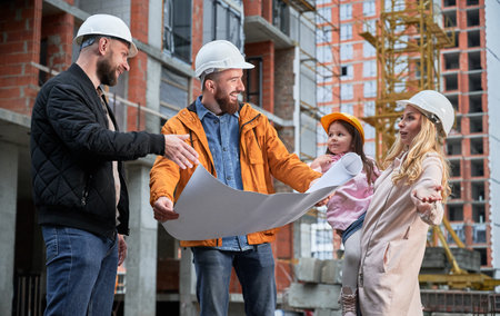 Happy family with child discussing architectural building plan with specialist at construction site. Parents with child and male builder standing outdoors near building under construction.の写真素材