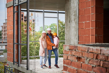 Full length of cheerful man holding daughter and smiling while standing next to wife at construction site. Happy parents with child posing in apartment building under construction.の写真素材