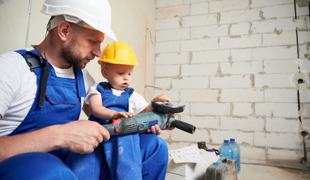 Little boy sitting on father lap and holding electric power grinding machine. Man and kid in work overalls using electric power tool in apartment under renovation with brick wall on background.の写真素材