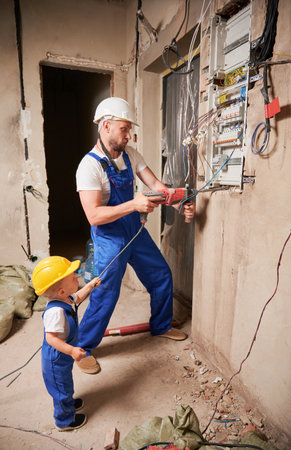 Male worker in safety helmet drilling wall with electric drill while little boy holding tool wire. Man and kid in work overalls installing electrical switch panel in apartment under renovation.の写真素材
