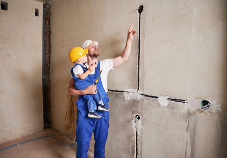 Male electrician in safety helmet holding little boy and pointing at electrical wire. Man and kid in work overalls standing by the wall and looking at electric cable in apartment under renovation.の写真素材