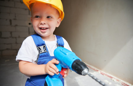 Cute baby boy construction worker looking aside and smiling while holding toy electric drill. Kid...