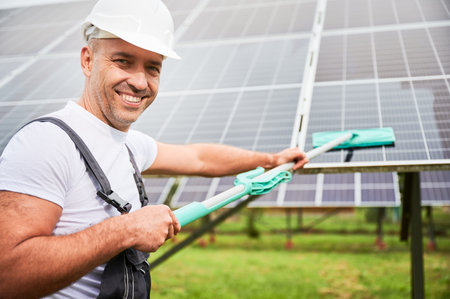 Smiling man wiping PV battery using mop. Professional worker doing good job at cleaning solar panel from dust. Man holding mop and smiling straight to camera.の写真素材