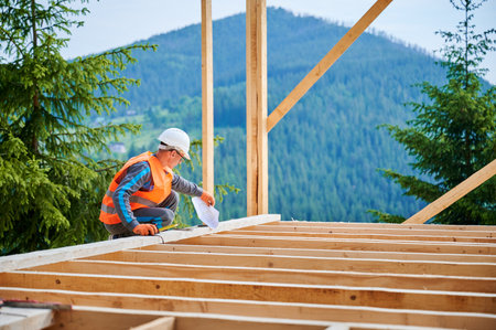 Carpenter in the process of building wooden frame house. Man measures distances with a measuring tape while dressed in workwear and helmet. Concept of modern, environmentally-friendly construction.の写真素材