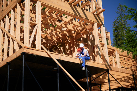 Father with toddler son building wooden frame house. Male builder sitting on the edge of the balcony at construction site, wearing helmets and blue overalls on sunny day. Carpentry and family concept.の写真素材