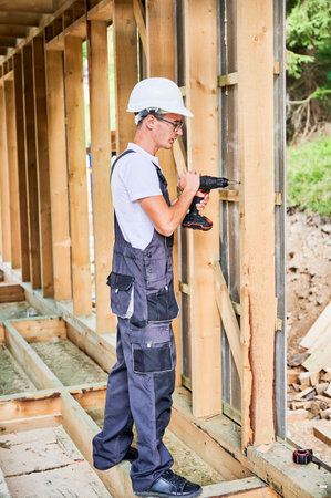 Carpenter constructing wooden framed house. Man worker in glasses working with screwdriver, wearing work overalls and helmet. Concept of modern eco-friendly construction.の写真素材