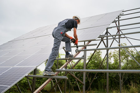 Worker fixing metal beams with electric drill for solar panels. Man wearing helmet and workwear. Photo-voltaic collection of modules as a PV panel. Array as a system of photo-voltaic panels.の写真素材