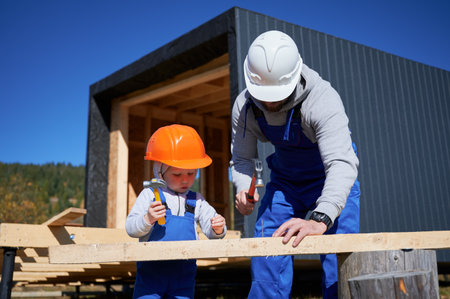 Father with toddler son building wooden frame house. Boy helping his daddy, hammering nail into plank on construction site, wearing helmet and blue overalls on sunny day. Carpentry and family concept.の写真素材