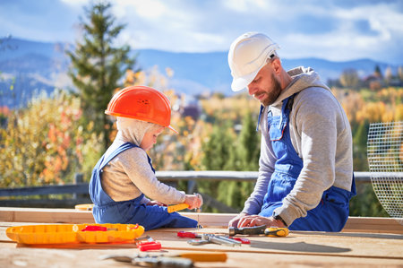 Father with toddler son building wooden frame house. Boy helping his daddy, hammering nail into plank on construction site, wearing helmet and blue overalls on sunny day. Carpentry and family concept.の写真素材