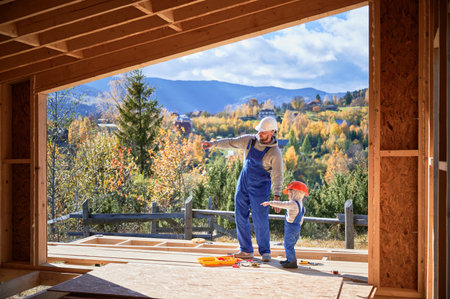 Father with toddler son building wooden frame house. Male builder with kid pointing on the wall on construction site, wearing helmet and blue overalls on sunny day. Carpentry and family concept.の写真素材