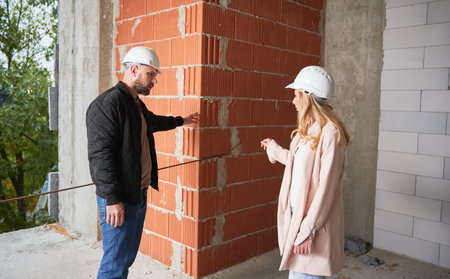 Woman standing by brick wall and discussing apartment replanning and renovation with specialist. Female homeowner having meeting with builder in building under construction.の写真素材
