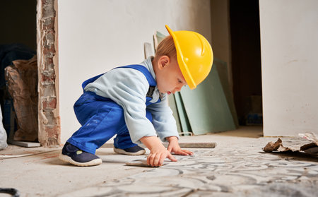 Little boy construction worker installing ceramic tile on the floor in apartment under renovation. Kid wearing safety helmet and work overalls while playing at home.の写真素材
