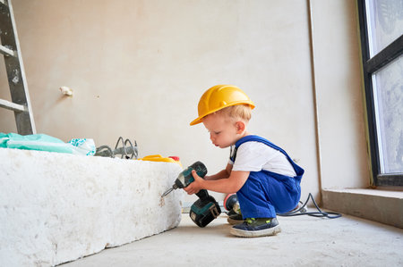 Adorable child in work overalls using electric construction tool in apartment under renovation. Cute kid in safety helmet working with power drill at home during renovation.の写真素材