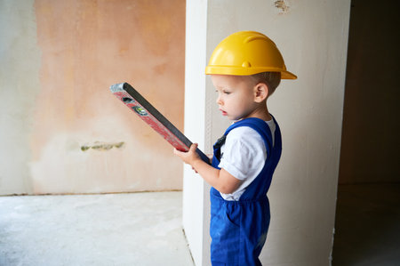 Child checking wall surface with spirit level in apartment. Kid in work overalls and safety construction helmet using level instrument while working on home renovation.の写真素材