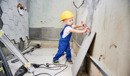 Child in safety construction helmet checking wall surface with spirit level in apartment. Kid in work overalls using level instrument while working on home renovation.の写真素材