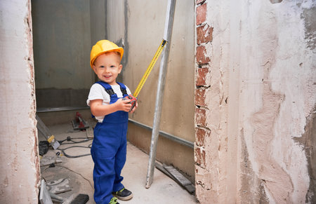 Happy boy construction worker holding measuring tape and smiling while standing in apartment under renovation. Cheerful kid with measuring tool wearing safety helmet and work overalls.の写真素材