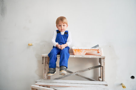 Cute kid construction worker sitting on wooden table against white wall in apartment under renovation. Cheerful little boy wearing safety helmet and work overalls while playing at home.の写真素材