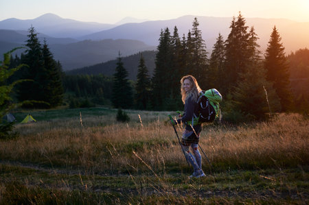Woman tourist hiking outdoors at sunset. Sporty, positive woman traveling in mountains. Young, sporty female with backpack using trekking sticks, walking, smiling. Concept of harmony with nature.の写真素材