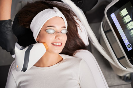 Top view of cheerful woman in safety goggles receiving laser facial treatment in cosmetology clinic. Beautician using diode laser scanner device while performing resurfacing skincare procedure.の写真素材