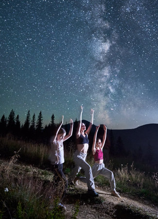 Three girls doing yoga at starry night. Sporty girls doing gymnastics with view of Milky Way. Yoga exercises in mountains.の写真素材