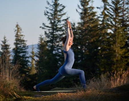 Sporty female exercising outdoor. Young woman doing warrior one pose. Workout on fresh air with view of mountains.の写真素材