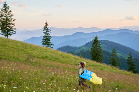 Happy Ukrainian running in Carpathian mountains. Little kid smiling and flattering Ukrainian flag on background of hills at sunset.の写真素材