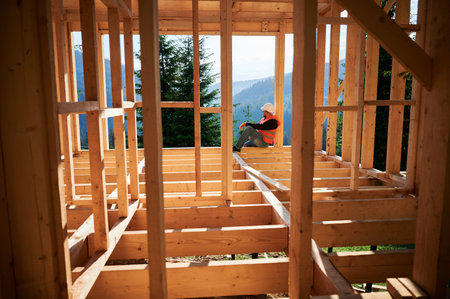 Carpenter constructing wooden frame house near the forest. Bearded man in glasses holding hammer, dressed in protective helmet and orange safety vest. Concept of ecological modern construction.の写真素材