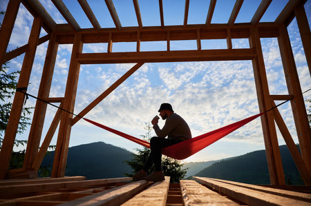 Man on construction site of wooden-framed house sits in hammock against backdrop of mountains and sunrise, sipping coffee. The concept pertains to modern ecological construction.の写真素材