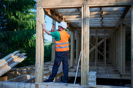 Carpenter constructing two-story wooden frame house near the forest. Man hammering nails into...