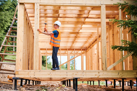 Wooden frame house being built by carpenter. Man wearing glasses hammering nails using hammer and is...