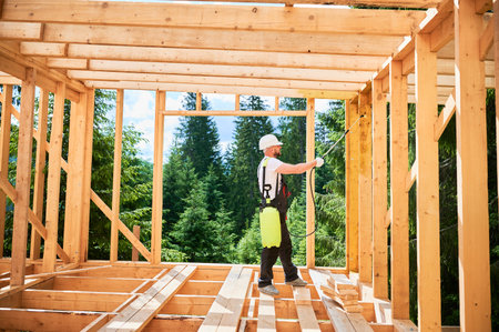 Laborer constructing wooden frame house near forest. Man treating woods, applying fire retardant using sprayer, while dressed in protective suit, helmet. Concept of modern eco-friendly construction.の写真素材