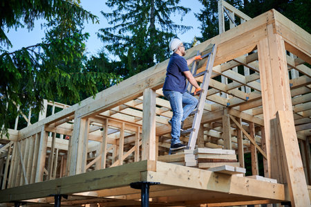 Carpenter constructing wooden frame house near the forest. Bearded man with glasses climbing ladder...
