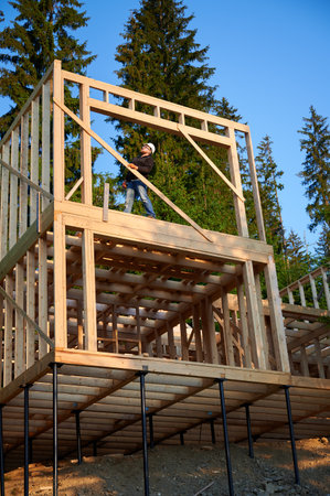 Carpenter constructing wooden framed two-story house near the forest. Bearded man with glasses holds...