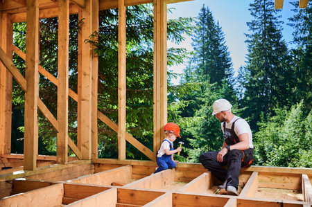 Father with toddler son building wooden frame house near forest. Male builder teaching his son the technique of pounding nails with hammer on construction site, wearing helmets and overalls.の写真素材