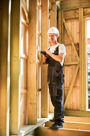 Carpenter constructing wooden framed house. Man worker working with screwdriver, wearing work overalls and helmet. Concept of modern eco-friendly construction.の写真素材