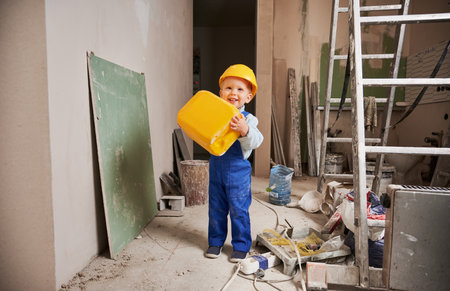 Cheerful little boy builder holding yellow construction material and smiling. Full length of adorable kid in safety helmet and work overalls standing in room with ladder. Home renovation concept.の写真素材