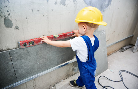 Child construction worker checking wall surface with spirit level tool in apartment. Kid in work overalls using level instrument while preparing wall for repair works at home during renovation.の写真素材