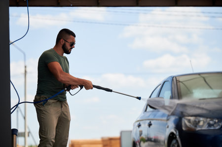 Man cleaning his car at self-service car wash with high pressure water jet. Confident man washing his blue auto with water gun at sunny day.の写真素材