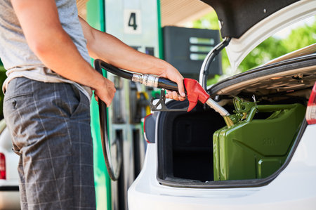 Male driver with gas pump refueling canister with fuel in car trunk. Man holding hose and filling cistern with gasoline. Close view of man filling canister on background of gasoline column.の写真素材