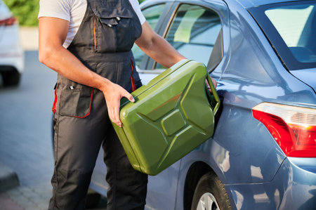 Stalled car filling with gasoline by professional worker. Young man refueling gas tank with fuel from canister. Cropped view of male worker in overalls filling blue car.の写真素材