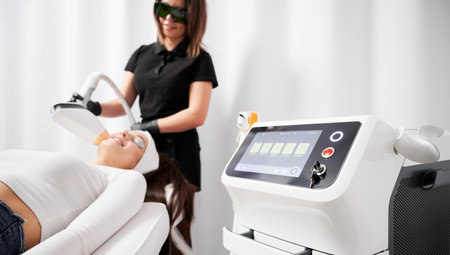 Selective focus of diode laser device cosmetology equipment with cosmetologist and patient on blurred background. Beautician performing laser skincare treatment in beauty clinic.の写真素材