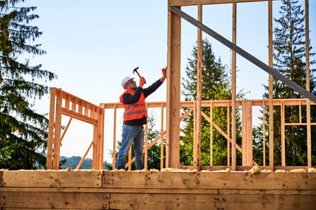 Wooden frame-built home is being erected by carpenter near forest. Bearded man wearing glasses driving in nails with hammer while sporting a safety helmet and a construction vest.の写真素材