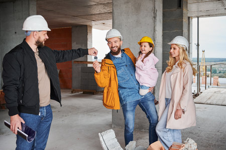 Male builder handing keys from new apartment to family with child at construction site. Happy...