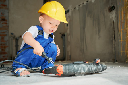 Cute child in safety helmet using rotary hammer drill in apartment under renovation. Kid construction worker playing with demolition drilling machine breaker tool. Home renovation concept.の写真素材