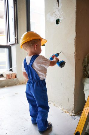 Boy in work overalls drilling wall with toy electric drill tool at home during renovation. Kid in safety helmet using drilling instrument while working on apartment refurbishment.の写真素材
